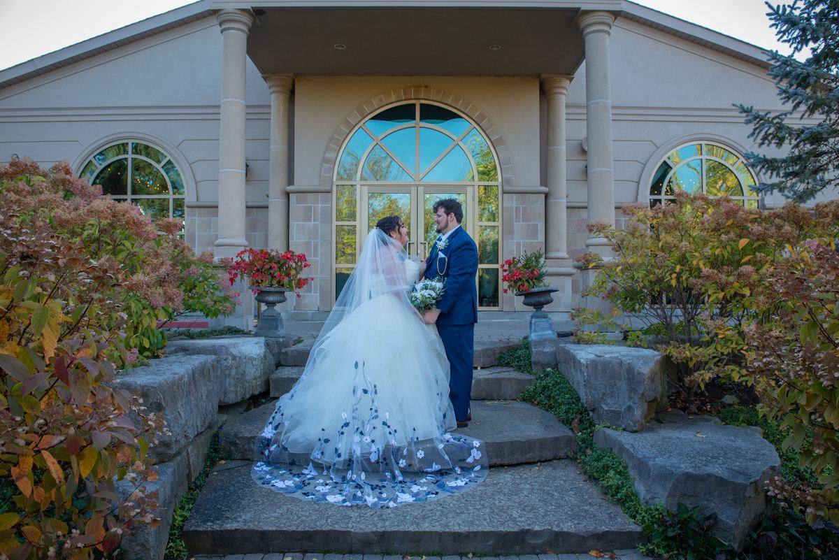 A bride and groom stand on stone steps, gazing at each other romantically as autumn leaves surround them.