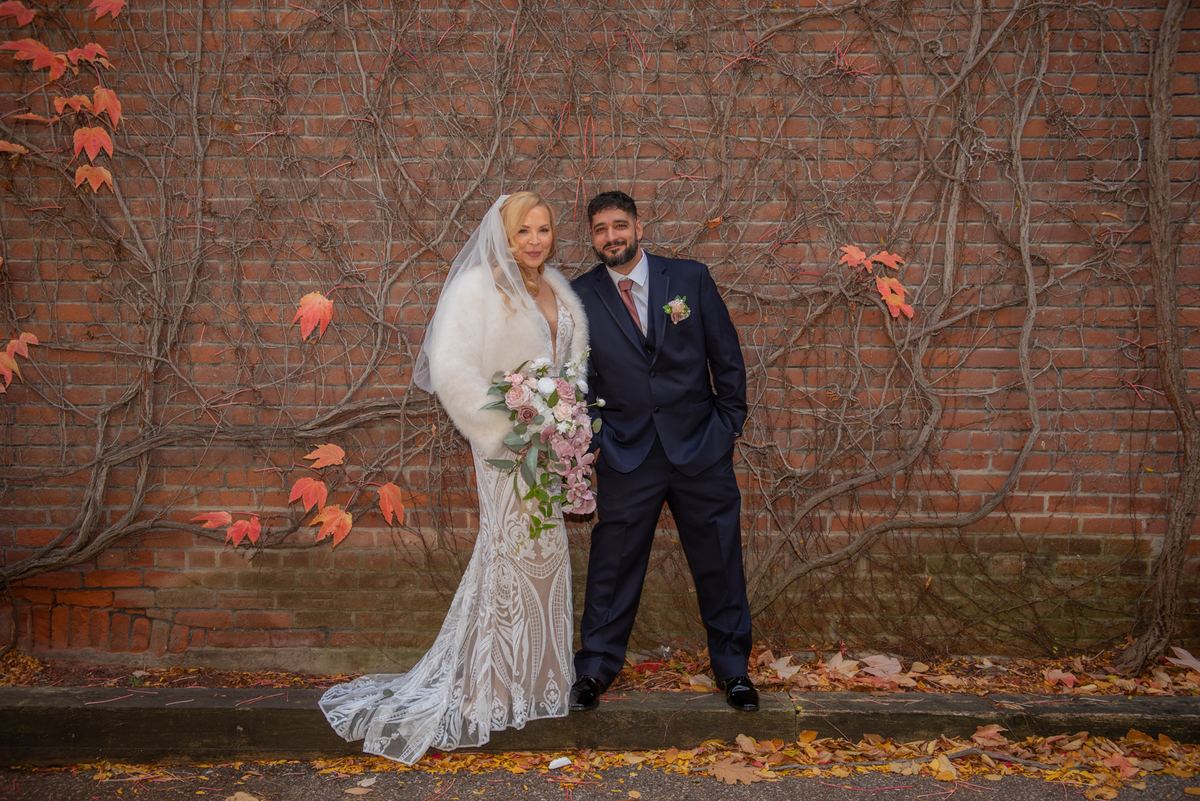 Bride in lace gown and fur stole with groom in navy suit stand by brick wall covered in autumn vines and leaves.
