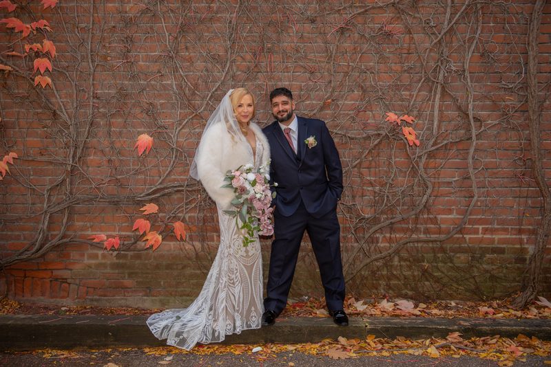 Bride in lace gown and fur stole with groom in navy suit stand by brick wall covered in autumn vines and leaves.