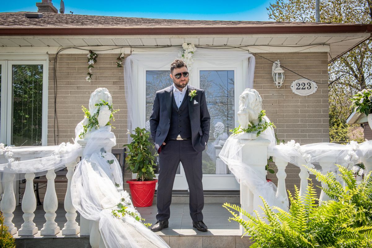 A groom stands on a decorated porch with two veiled statue brides, under bright daylight and festive greenery.