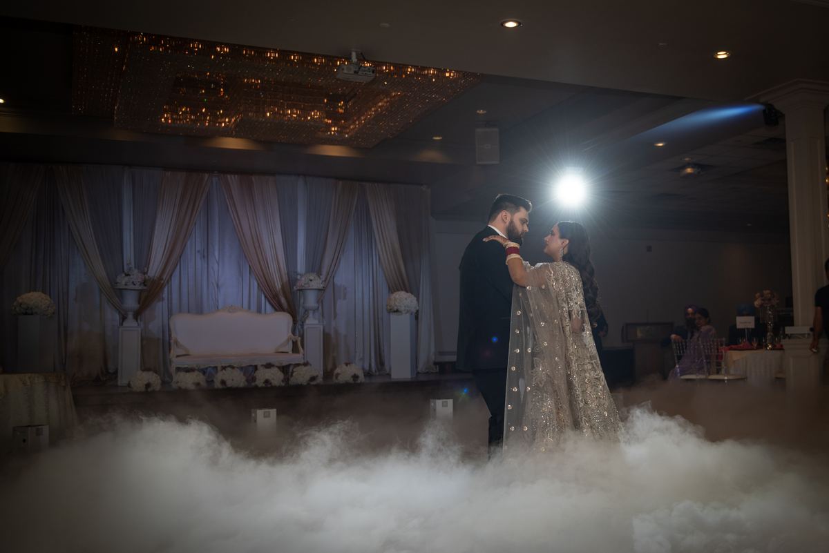 A newlywed couple dances intimately on a foggy stage, illuminated by a spotlight, gazing at each other.