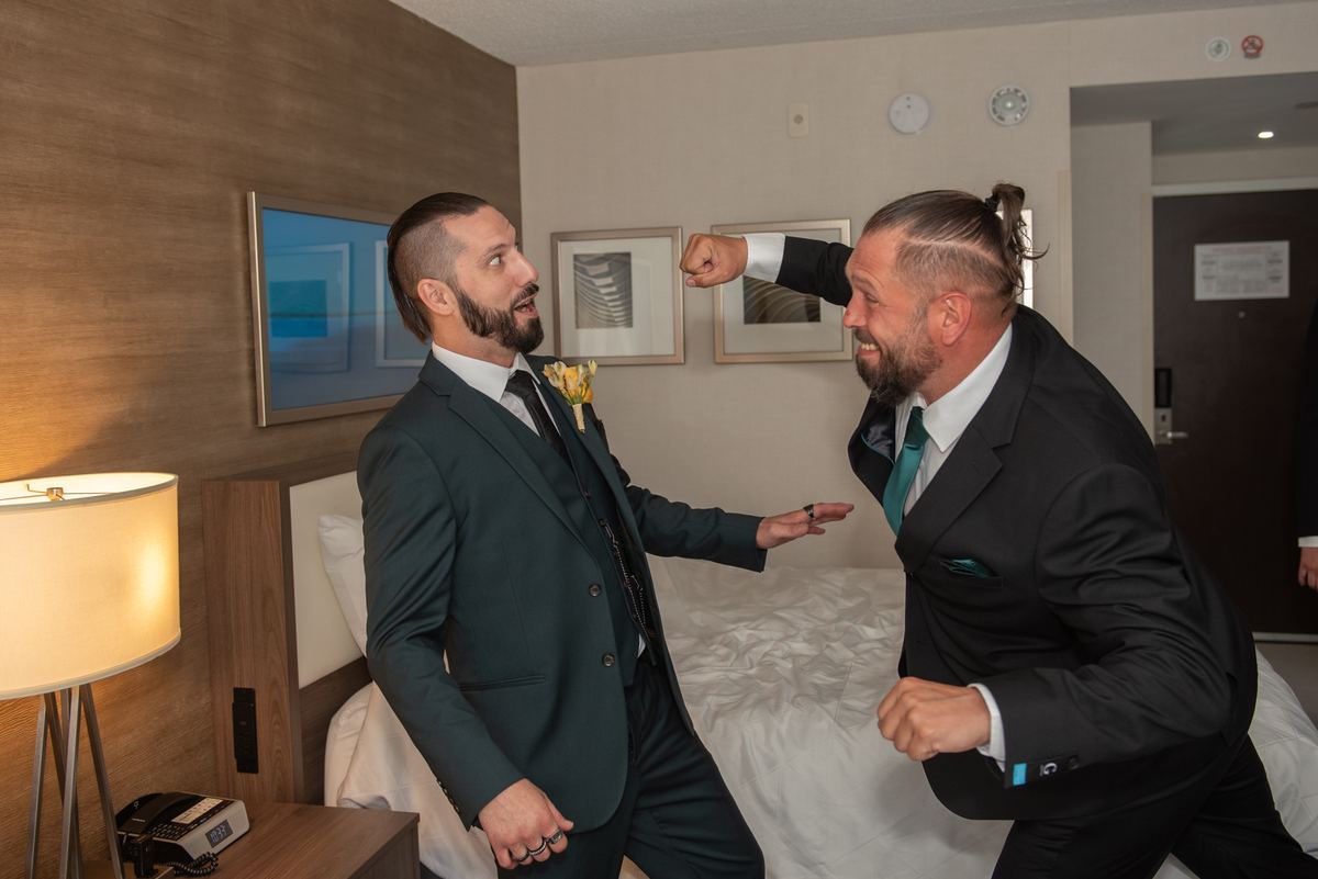 Two men in suits playfully engage in a mock fight, one extending a fist while the other gestures back.