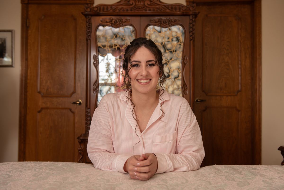 A bride smiles at the camera in a cozy wood-paneled room, wearing a pink pajama top and sitting by an ornate window.