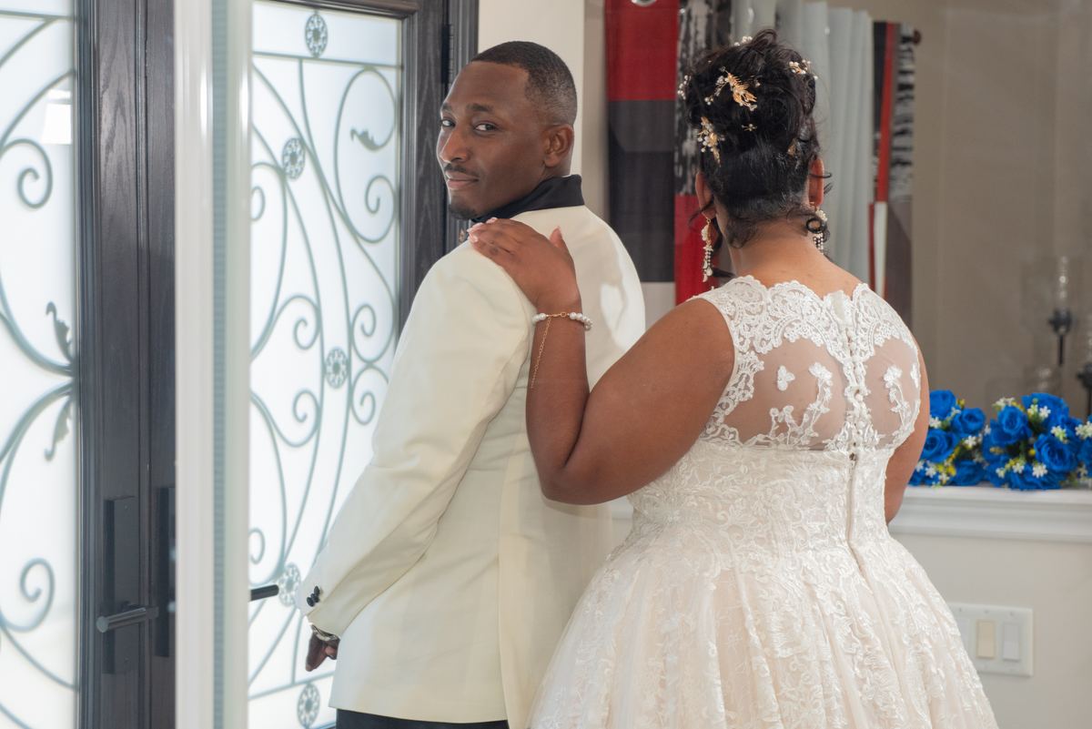 A bride and groom stand back-to-back in an indoor space with ornate glass doors and blue floral arrangements.