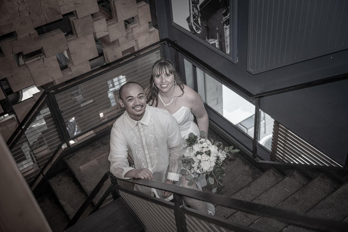 A bride and groom smile from a modern staircase, the scene in monochrome with natural light highlighting wood.