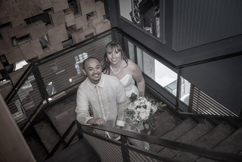A bride and groom smile from a modern staircase, the scene in monochrome with natural light highlighting wood.