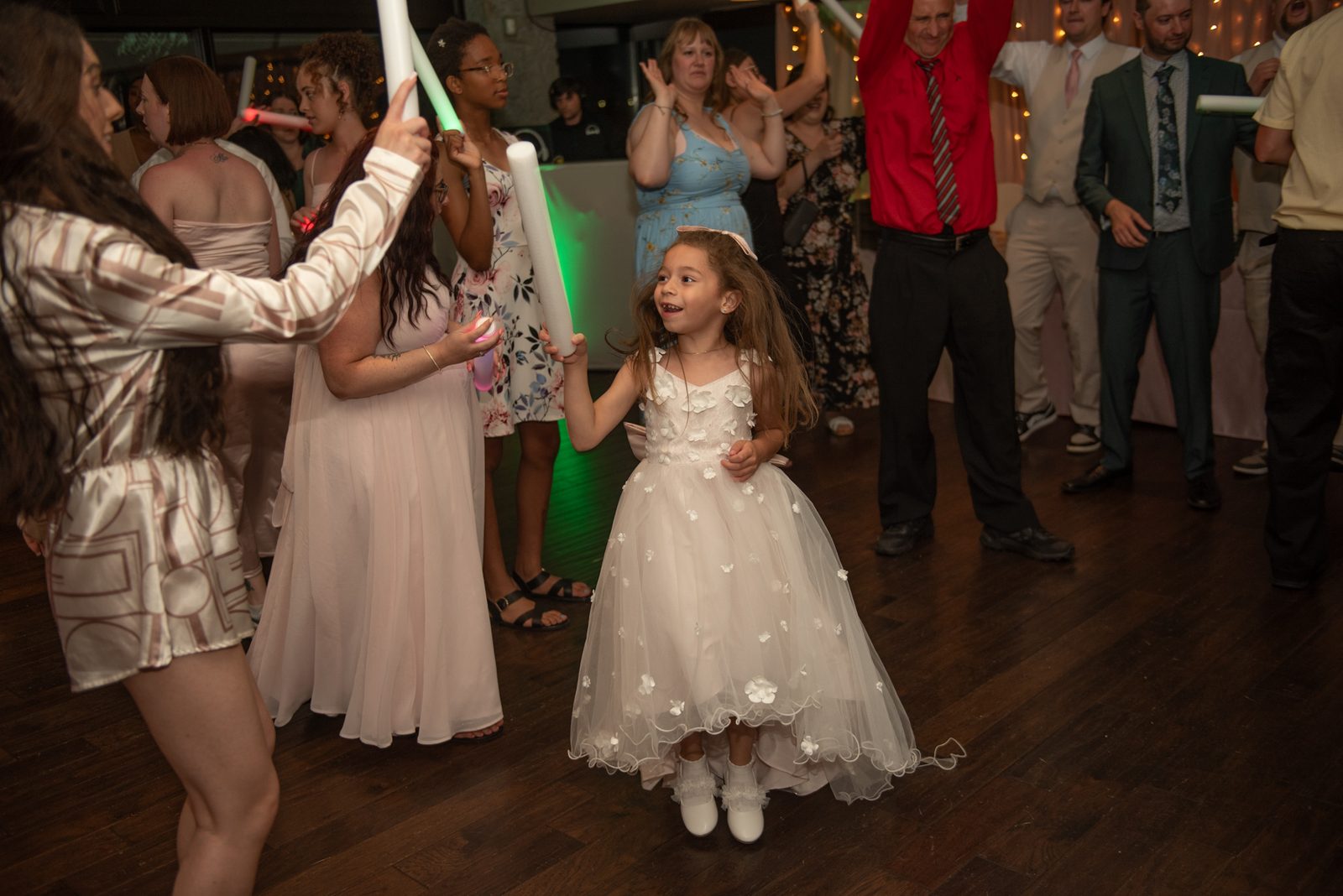 A girl in a white floral dress dances with a green light stick as others clap and hold similar sticks.