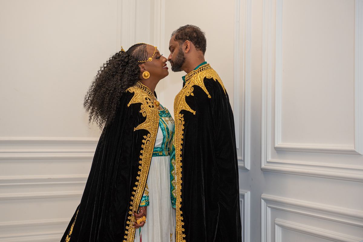 A couple in black velvet robes with gold embroidery stand close, foreheads nearly touching, against a plain white wall.