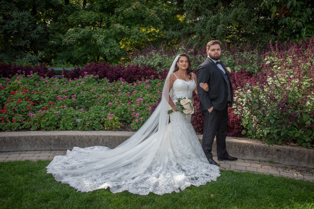 A bride in a lace gown and veil stands with her groom in a dark suit in a sunlit garden, surrounded by trees.