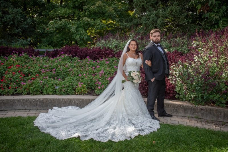 A bride in a lace gown and veil stands with her groom in a dark suit in a sunlit garden, surrounded by trees.