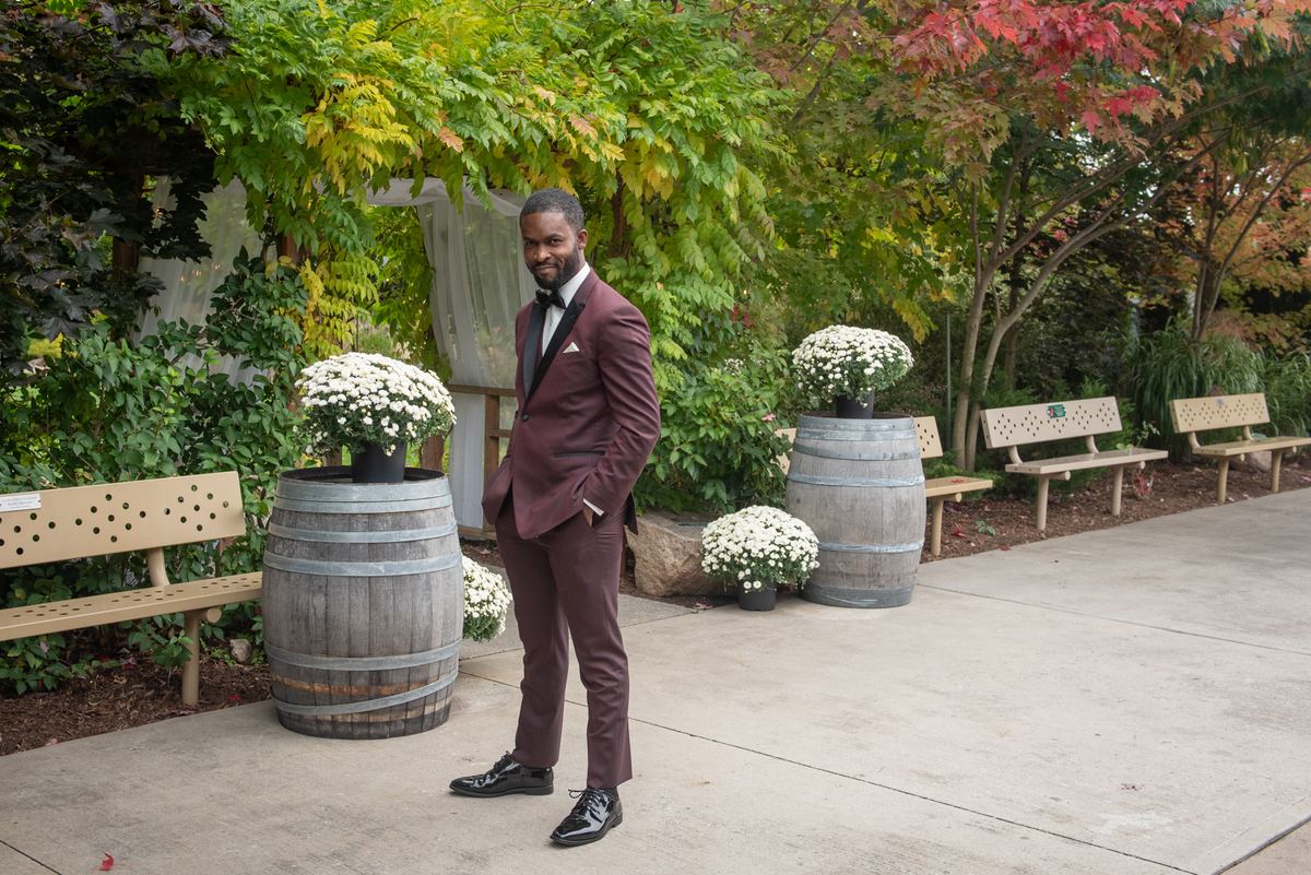 A groom in a burgundy tuxedo stands confidently among autumn trees and greenery, smiling at the camera.