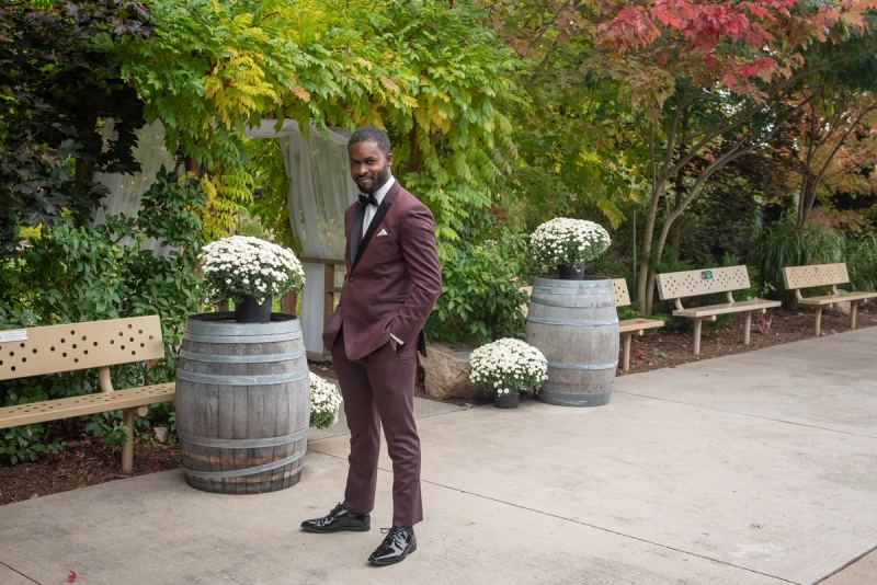 A groom in a burgundy tuxedo stands confidently among autumn trees and greenery, smiling at the camera.