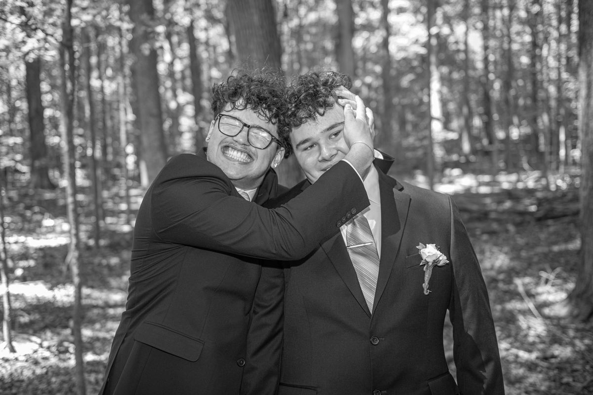 Two men in suits laugh and embrace playfully in a sun-dappled forest, highlighting their joyful connection.