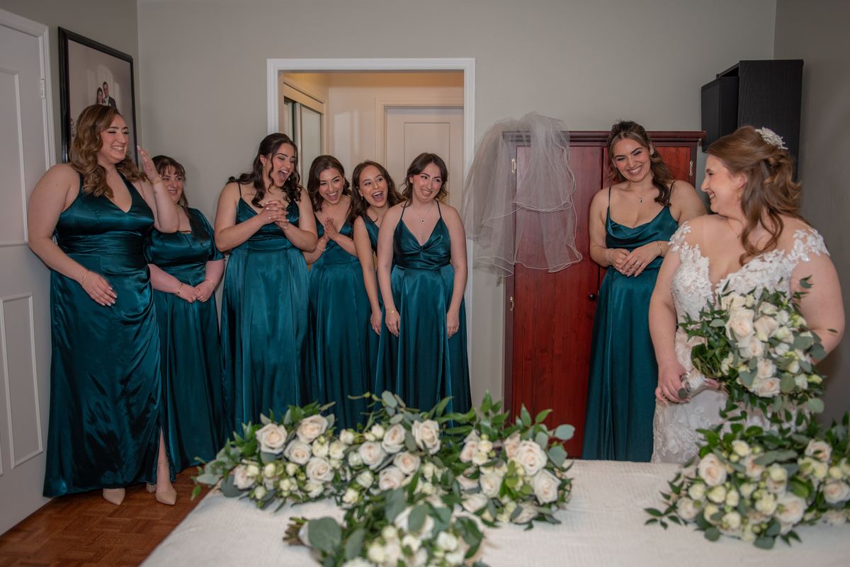 A bride in a white lace gown stands with her smiling bridesmaids in teal satin dresses near a doorway.
