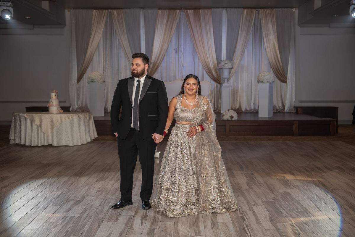 A bride in a shimmering lehenga and groom in a tuxedo stand on a wooden floor, smiling at the camera under a spotlight.
