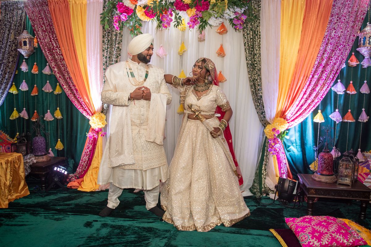 A South Asian bride and groom smile warmly under a floral canopy adorned with colorful tassels and rich fabrics.