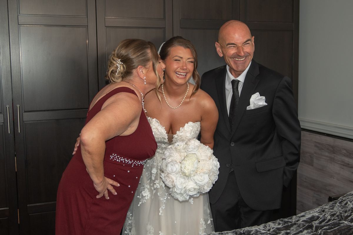 A bride laughs as a woman in maroon whispers, with the groom smiling beside them, near a dark wooden door.