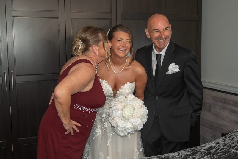 A bride laughs as a woman in maroon whispers, with the groom smiling beside them, near a dark wooden door.