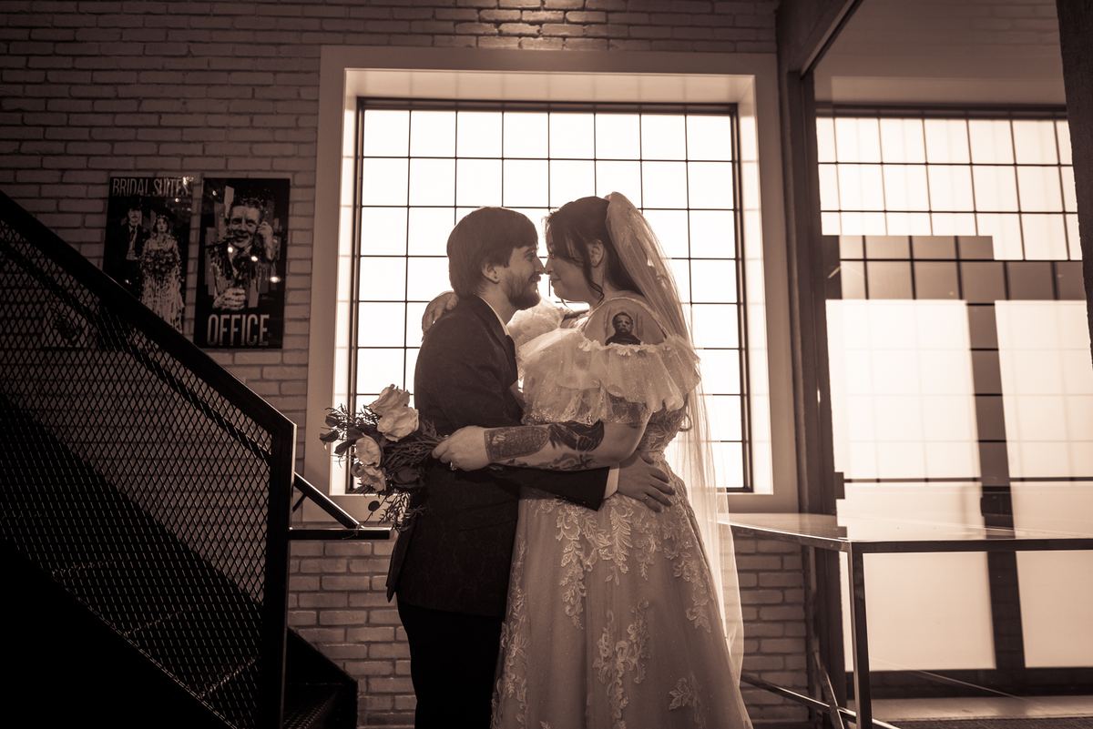 A newlywed couple embraces in a sepia-toned room with large windows and brick walls, holding a bouquet near a staircase.
