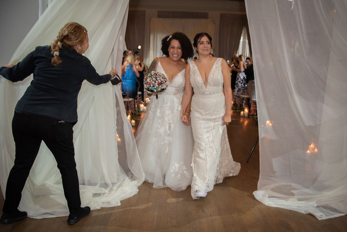 Two brides walk down a candlelit aisle, framed by white drapes parted by an attendant, in a warmly lit ceremony.