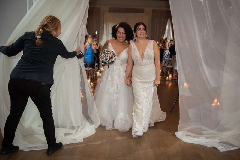 Two brides walk down a candlelit aisle, framed by white drapes parted by an attendant, in a warmly lit ceremony.