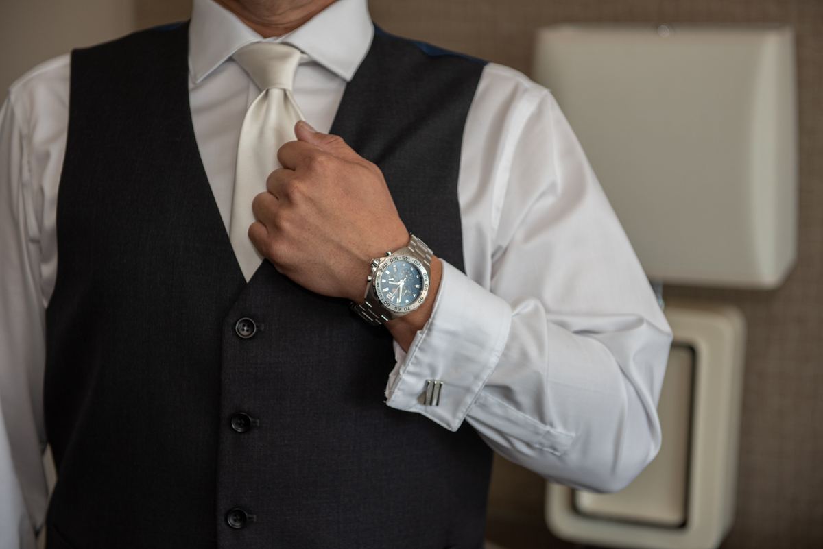 A man in formal attire adjusts his tie while wearing a silver watch, preparing for an event in a softly lit room.