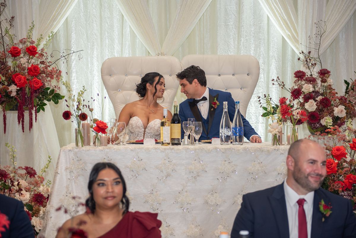 A bride and groom smile at each other warmly at a lavishly decorated reception table, surrounded by guests in formal attire.