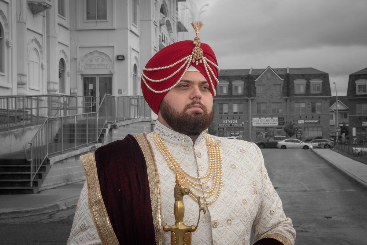 A man in traditional Indian wedding attire stands outdoors against an urban backdrop on a cloudy day.