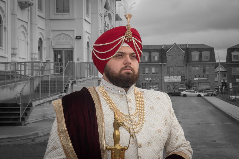 A man in traditional Indian wedding attire stands outdoors against an urban backdrop on a cloudy day.