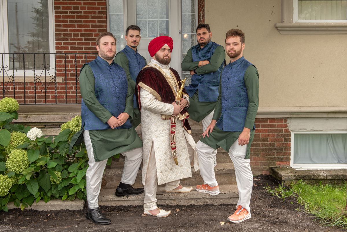 A groom in white and red stands with four groomsman in blue and green vests, framed by hydrangeas under soft daylight.