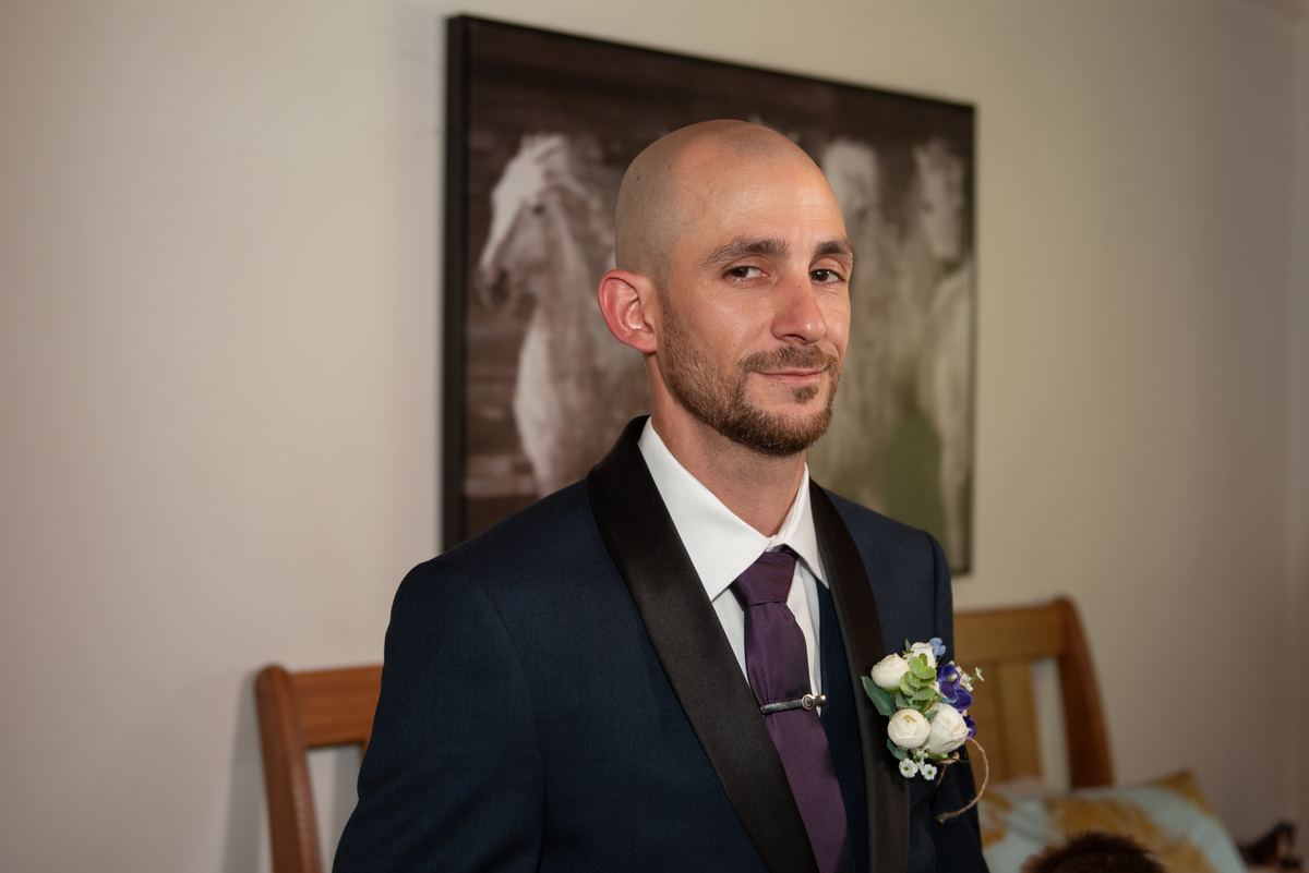 A groom in a navy tuxedo with a purple tie and boutonnière looks tenderly off-camera near a wooden chair.