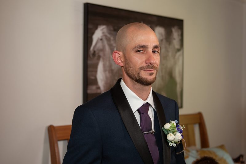 A groom in a navy tuxedo with a purple tie and boutonnière looks tenderly off-camera near a wooden chair.