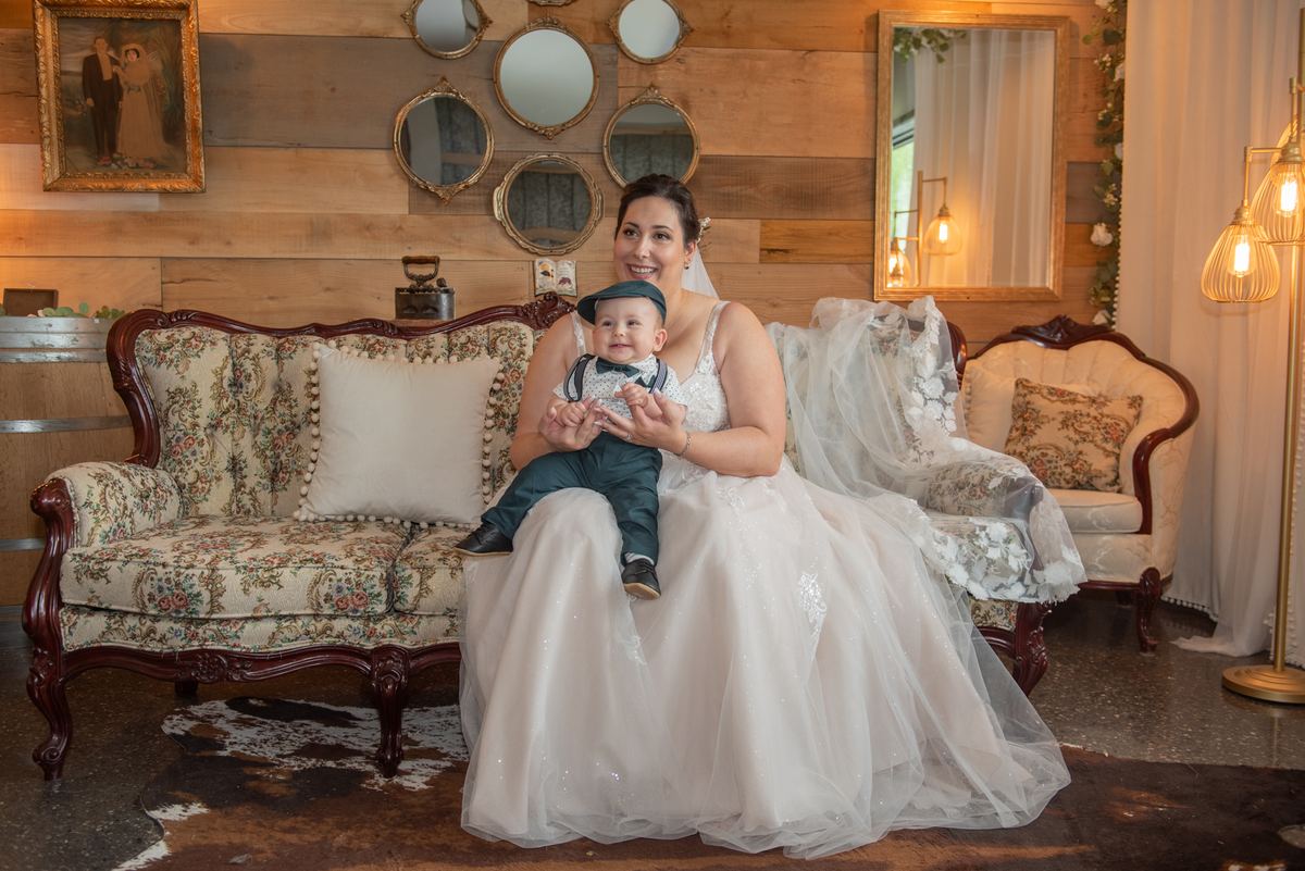 A bride in a white gown sits with a child in a miniature suit on an ornate floral sofa, both smiling at the camera.