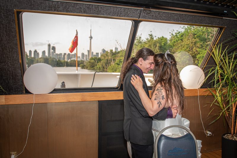 A newlywed couple embraces joyfully in a yacht cabin, gazing at Toronto's skyline through the window.