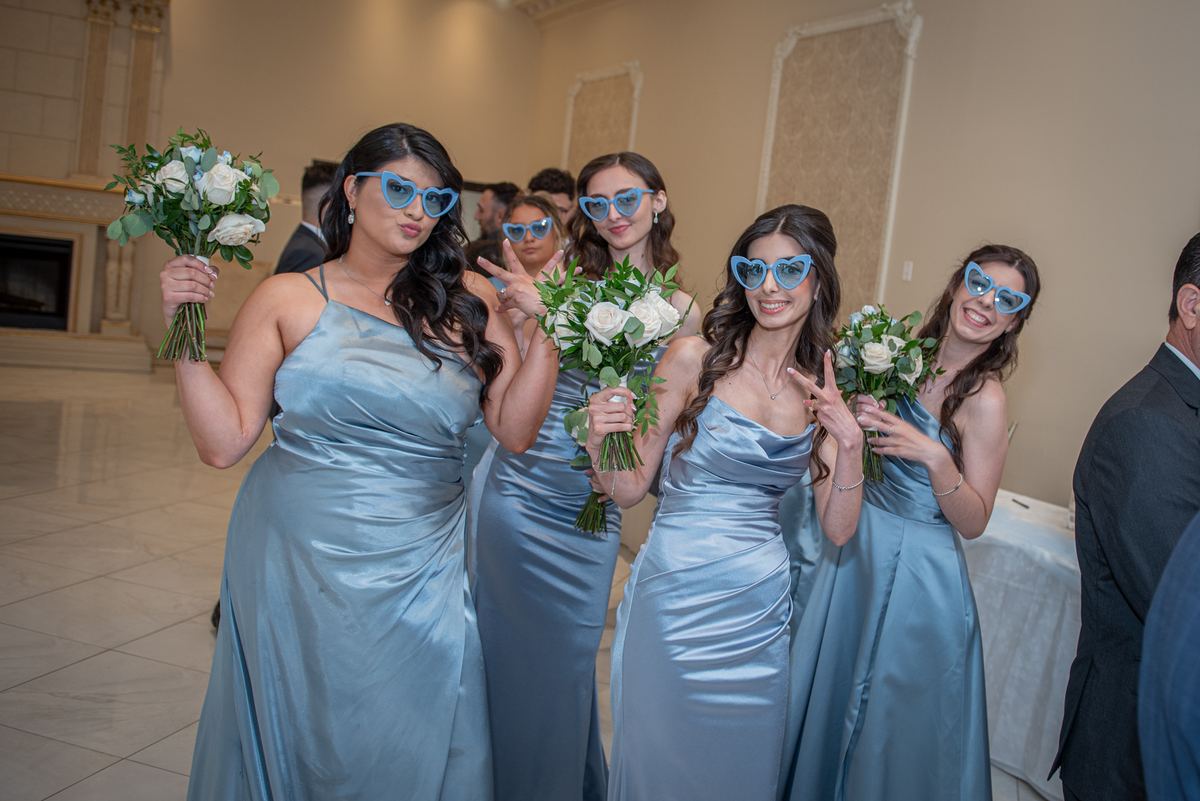 Bridesmaids in light blue gowns pose playfully with heart-shaped sunglasses and white roses indoors.