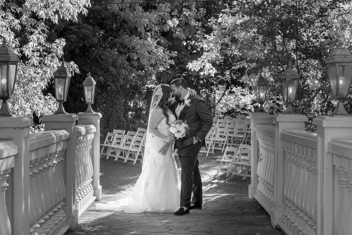 A newlywed couple kisses on a stone walkway with white railings and lanterns, under dappled sunlight from trees.