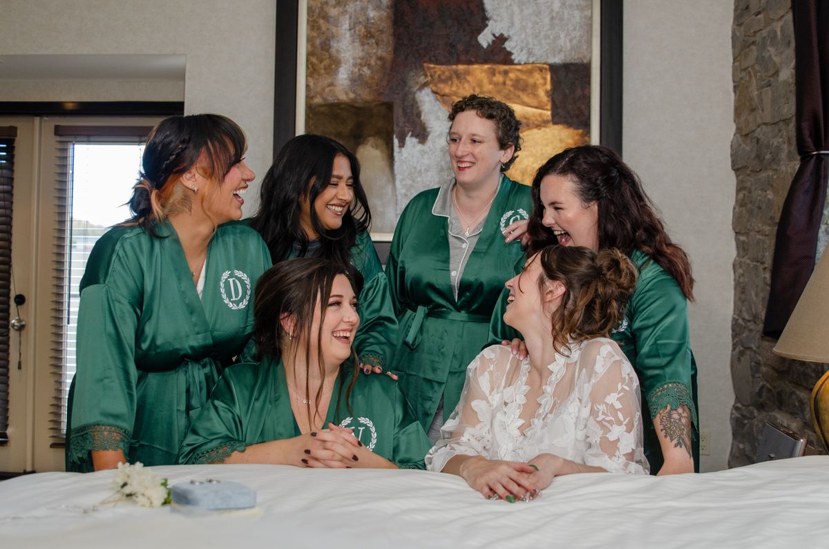A bride in a white lace gown sits with her laughing bridesmaids in green satin robes on a bed, surrounded by warmth.