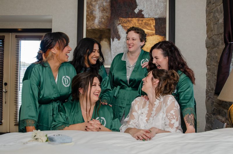 A bride in a white lace gown sits with her laughing bridesmaids in green satin robes on a bed, surrounded by warmth.