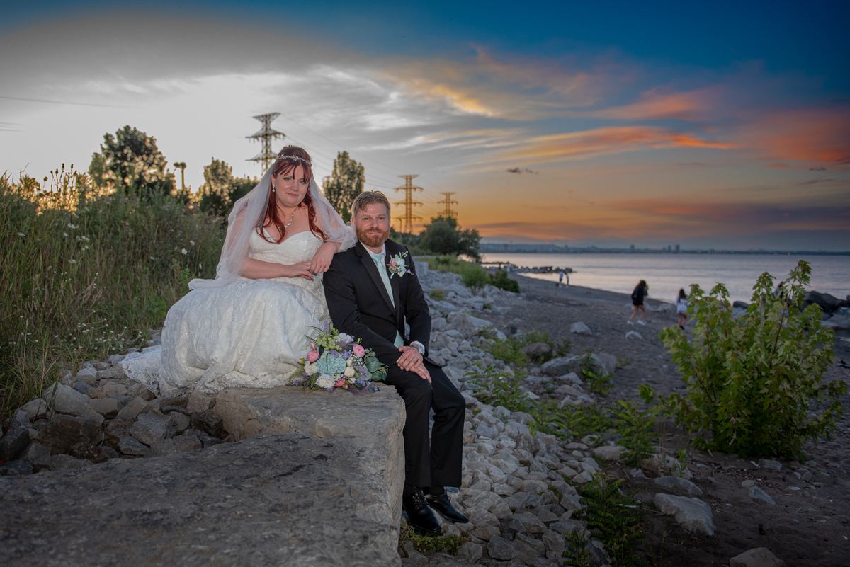 A bride and groom pose on a stone embankment by the waterfront at sunset, holding flowers and wearing formal attire.