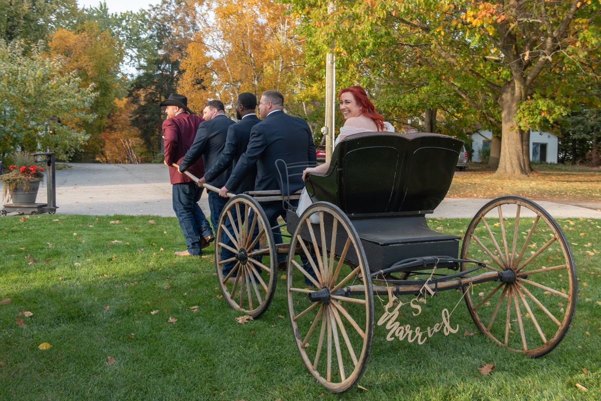 A smiling red-haired bride in a carriage with a 'Just Married' sign as four men push it across an autumn lawn.