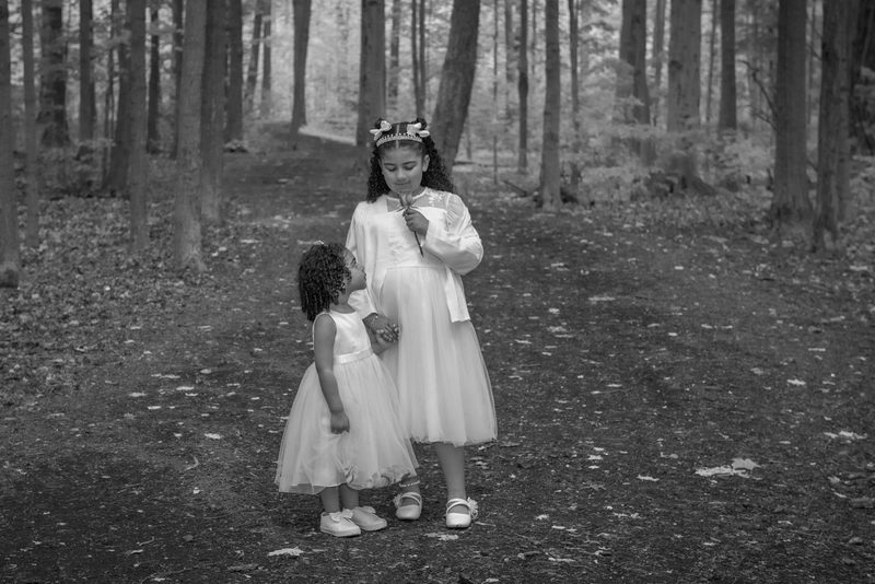 Two young girls in white dresses stand亲密换行符接近，一个女孩抬头仰望着另一个女孩，后者手拿一朵小花放在唇边。阳光透过树木洒在她们身上，营造出一种宁静而私密的氛围。