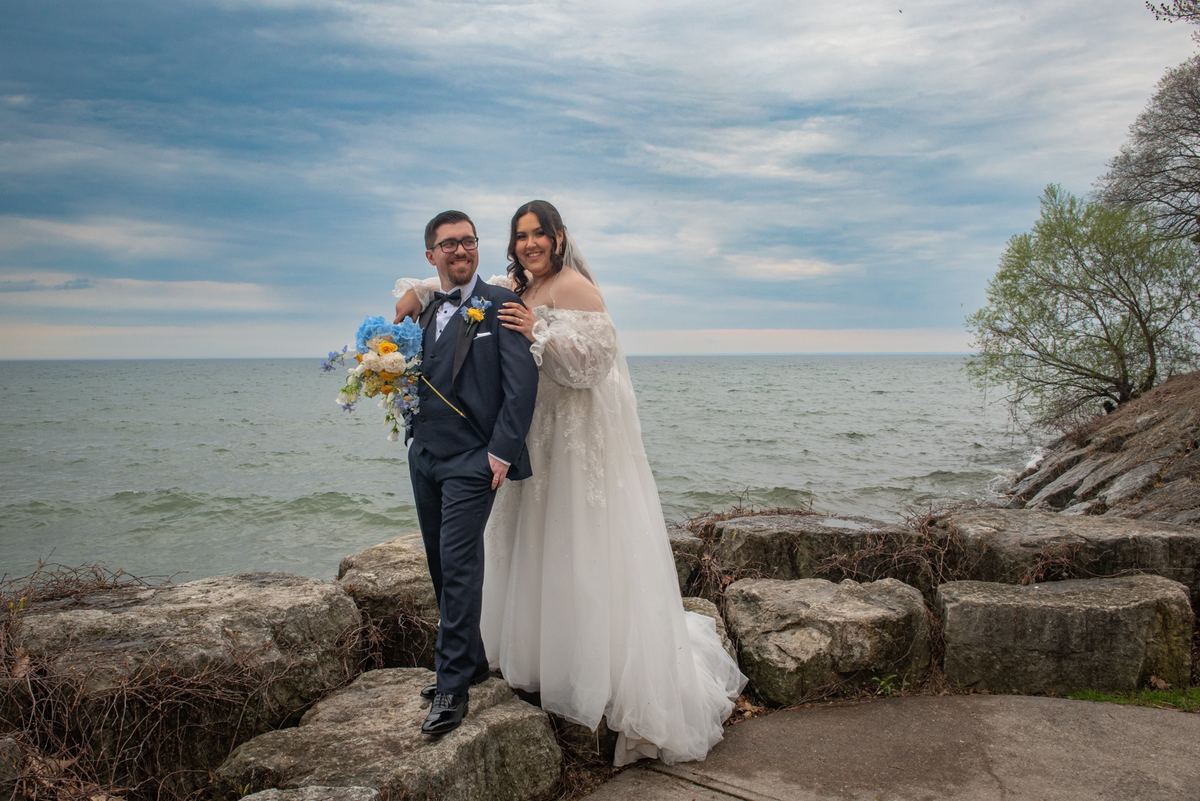 A newlywed couple poses by a lake, the groom in a dark suit holding flowers as the bride embraces him.