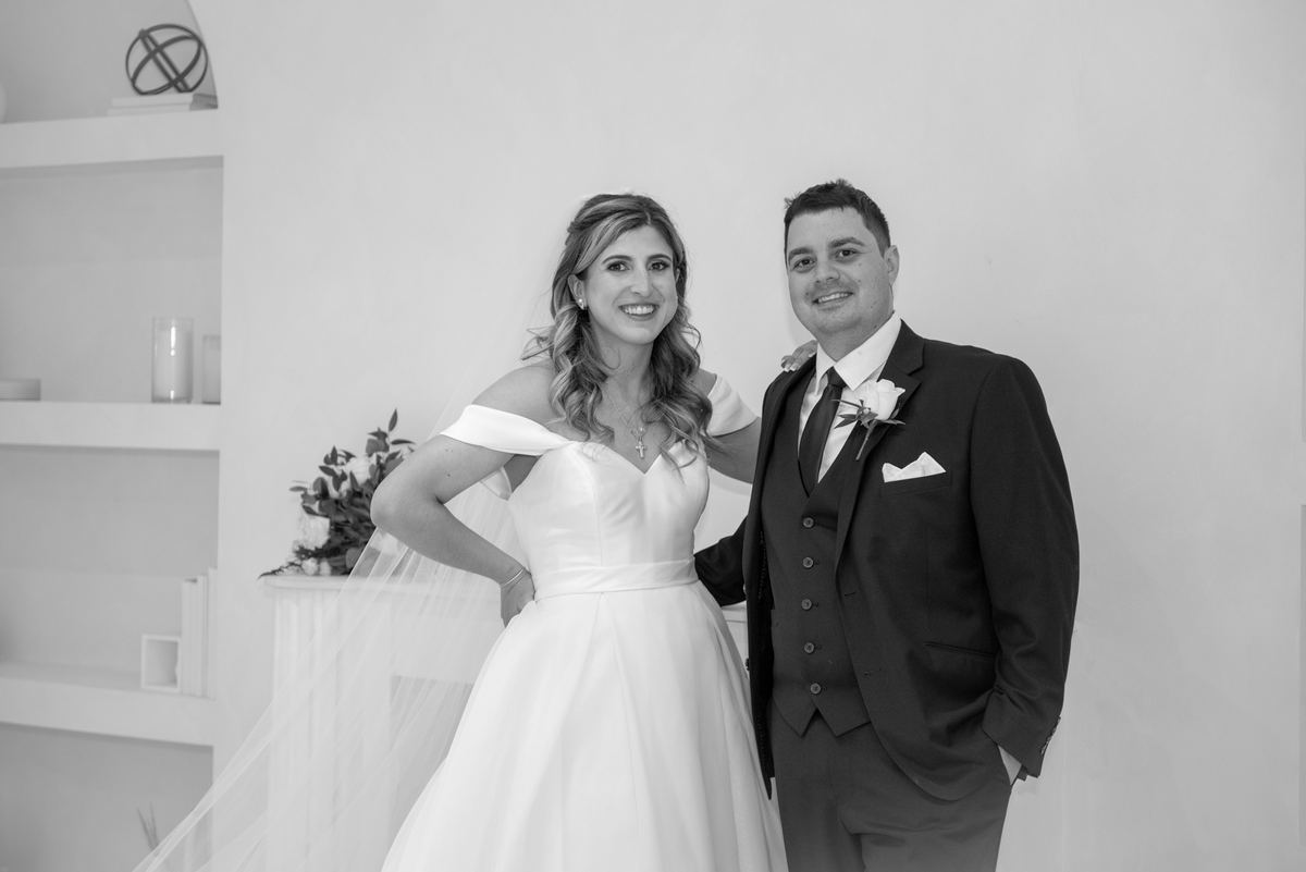 A smiling bride in an off-the-shoulder gown and veil stands confidently next to her groom in a dark suit.