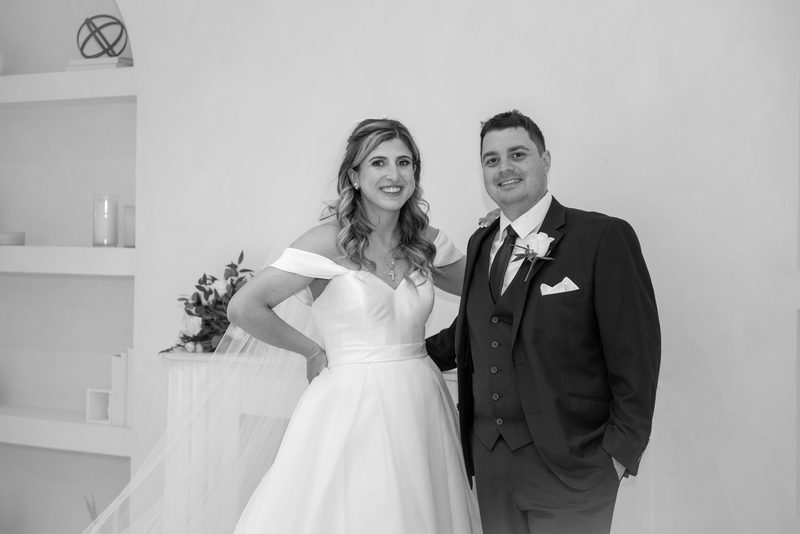 A smiling bride in an off-the-shoulder gown and veil stands confidently next to her groom in a dark suit.
