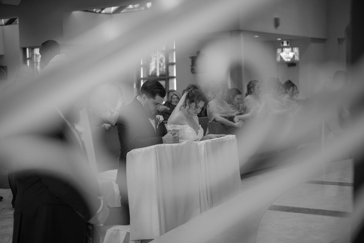 A bride and groom lean over a white-draped table during their wedding ceremony, focused on a task in soft light.