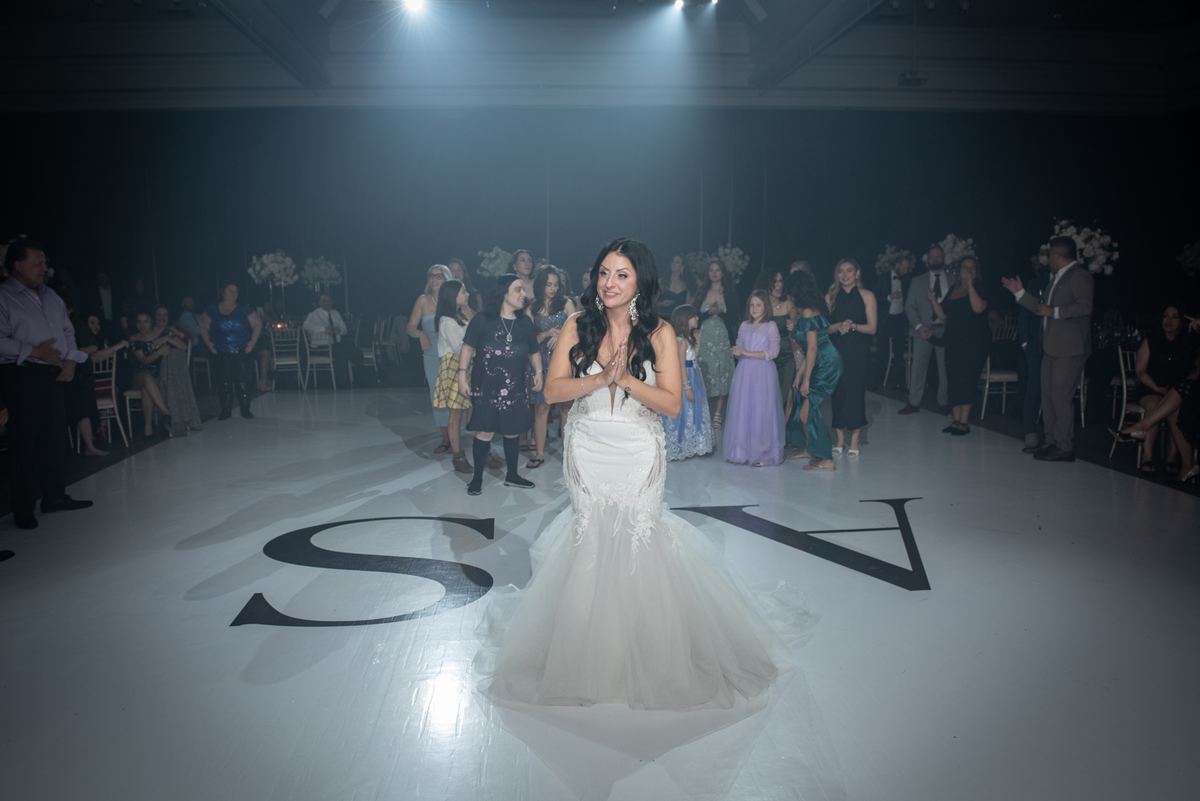 A bride in a white mermaid gown stands center stage under spotlights, hands clasped, as guests applaud from the periphery.