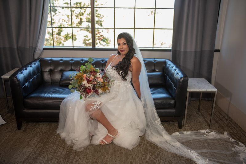 A bride in a white gown and veil sits on a black leather sofa, holding a bouquet and smiling at the camera.