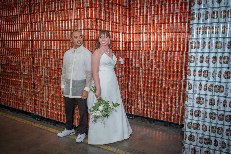 A bride and groom smile next to stacks of beverage cans in a warehouse setting, under bright lights.