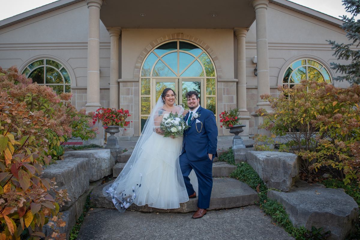 A bride and groom smile on stone steps outside an elegant building with autumn foliage, potted flowers, and warm sunlight.