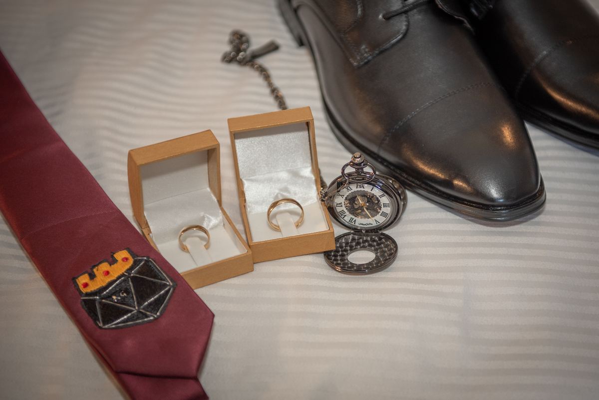 Two wedding rings lie in open boxes next to a vintage pocket watch and dress shoes, suggesting pre-wedding preparations.